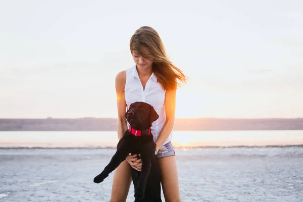 Jovem abraço feminino com cão labrador retriever na praia ao pôr do sol — Fotografia de Stock