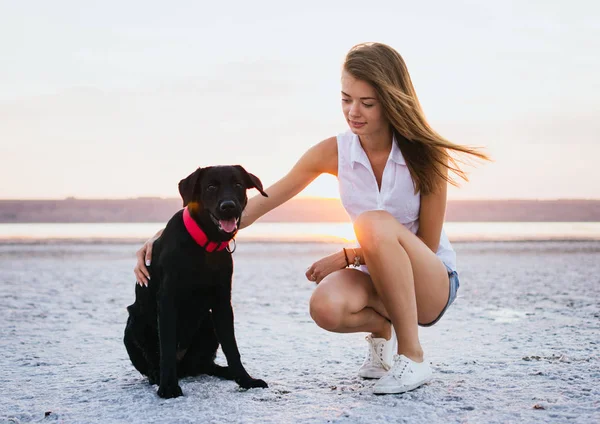 Jovem abraço feminino com cão labrador retriever na praia ao pôr do sol — Fotografia de Stock