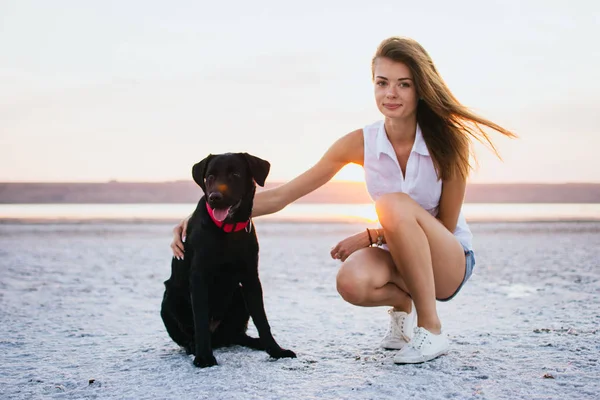 Jovem abraço feminino com cão labrador retriever na praia ao pôr do sol — Fotografia de Stock