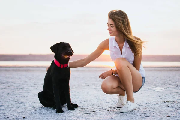 Jovem abraço feminino com cão labrador retriever na praia ao pôr do sol — Fotografia de Stock