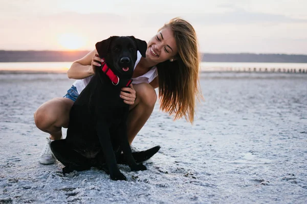 Šťastná mladá žena s pes labrador retriever na pláži při západu slunce — Stock fotografie