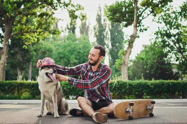 Giovane hipster seduto su skateboard con divertente cane husky siberiano in cap — Foto Stock
