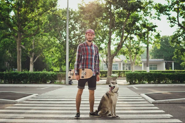 Junger Hipster-Mann mit Skateboard und sibirischem Husky-Hund steht auf Straße — Stockfoto