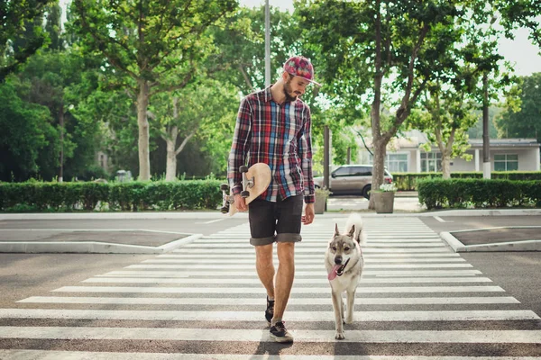Junger Hipster-Mann läuft mit Skateboard und sibirischem Husky-Hund auf Straße — Stockfoto