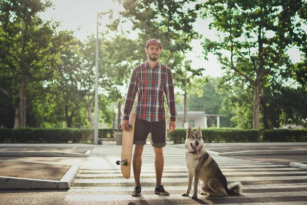Joven hipster hombre con monopatín y perro husky siberiano de pie en la calle — Foto de Stock