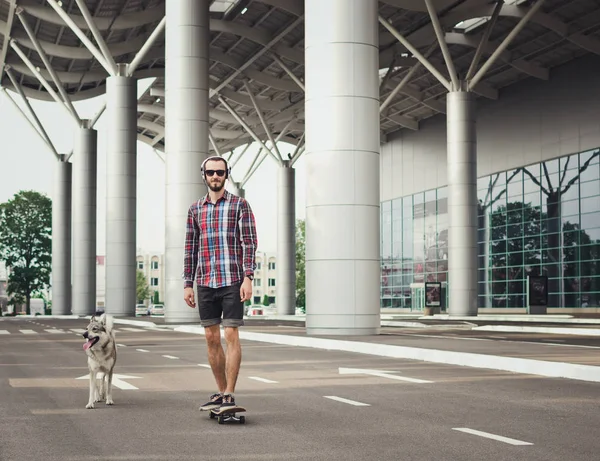 Joven hipster hombre paseos en el monopatín con perro husky en la calle carretera —  Fotos de Stock