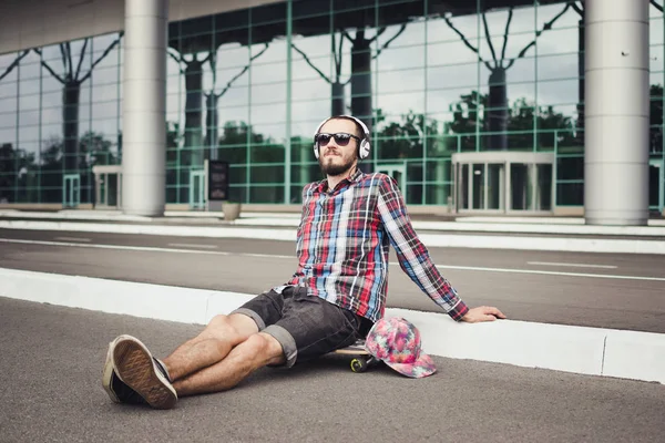 Porträt eines jungen Hipster-Mannes, der auf dem Skateboard auf der Straße sitzt und Musik hört — Stockfoto