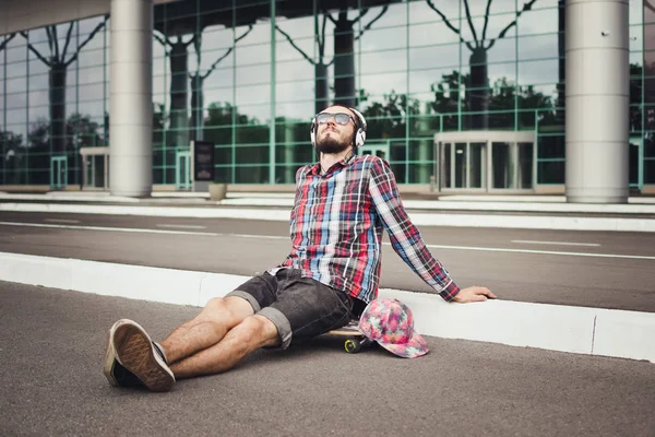 Retrato del joven hipster sentado en el monopatín en la calle y escuchar música — Foto de Stock