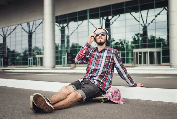 Retrato del joven hipster sentado en el monopatín en la calle y escuchar música — Foto de Stock