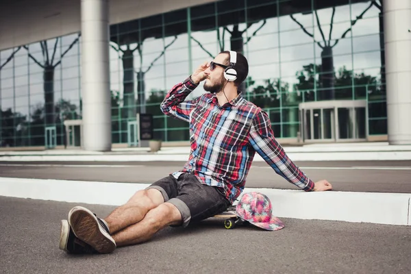 Retrato del joven hipster sentado en el monopatín en la calle y escuchar música — Foto de Stock