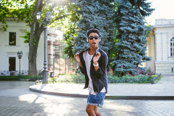 Retrato de un joven y elegante hombre negro hipster con auriculares blancos y gafas de sol bailando al aire libre en el centro de la ciudad, divirtiéndose — Foto de Stock