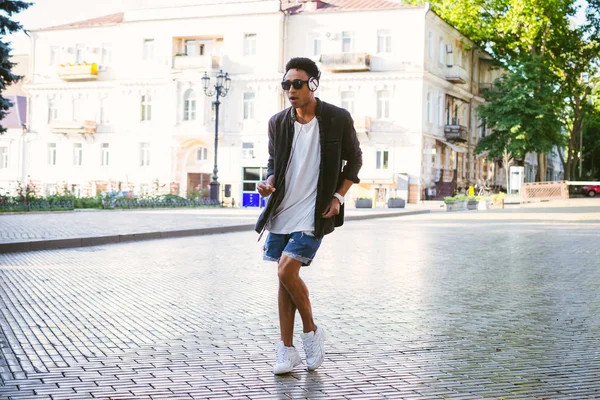 Retrato de jovem elegante homem negro hipster em fones de ouvido brancos e óculos de sol dançando ao ar livre no centro da cidade, se divertindo — Fotografia de Stock