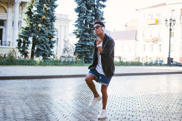 Retrato de jovem elegante homem negro hipster em fones de ouvido brancos e óculos de sol dançando ao ar livre no centro da cidade, se divertindo — Fotografia de Stock