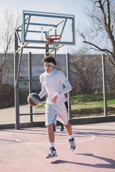 Retrato de um jogador de basquete andando em um campo de basquete ao ar livre e driblando a bola — Fotografia de Stock