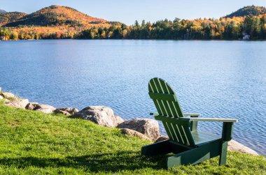 Güneşli bir günde bir göl kıyısında yeşil çimenlerin üzerinde boş Adirondack sandalye.