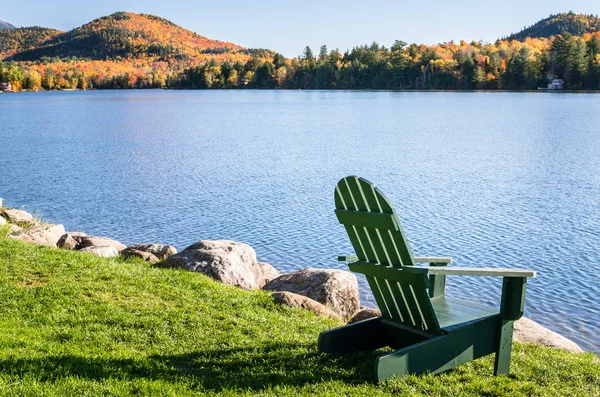 Güneşli bir günde bir göl kıyısında yeşil çimenlerin üzerinde boş Adirondack sandalye.