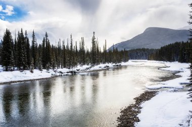Bulutlu bir kış gününde karla kaplı ormanlık bankalarla doğal Bow Nehri. Banff Ulusal Parkı, Ab, Kanada.