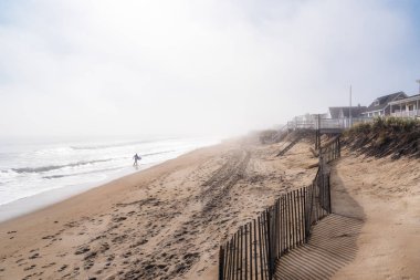 Yalnız sörfçü, bir sonbahar sabahı kalın bir sis tabakası altında sahil boyunca sörf tahtası taşıyor. Seabrook, Nh, Usa.