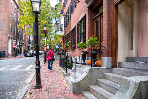 Lonely woman walking along a brick sidewalk lined traditional American townhouses on a cloudy autumn day. Beacon hill, Boston, MA, USA.