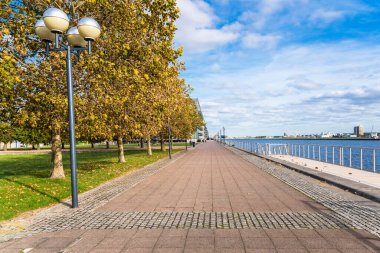 Parçalı bulutlu bir sonbahar gününde Londra rıhtımlarında ağaçlarla ve sokak ışıklarıyla kaplı bir nehir kenarı parkı. Londra, İngiltere.