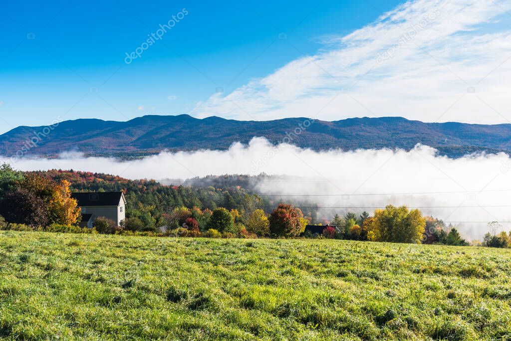 Mountain landscape with a patch of fog covering a forested valley on a fall morning. Beautiful autumn colours and blue sky. Waterbury, VT, USA.