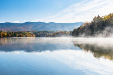 Majestic mountain lake with forested shores in autumn. The surface of the lake is covered in morning fog. Stunning autumn colours. Waterbury, VT, USA. clipart