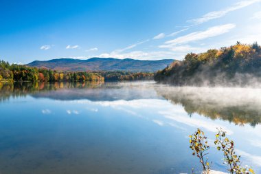 Beautiful lake with fog lifting from the water surface in a wooded mountain landscape on a sunny autumn morning. Waterbury, VT, USA. clipart