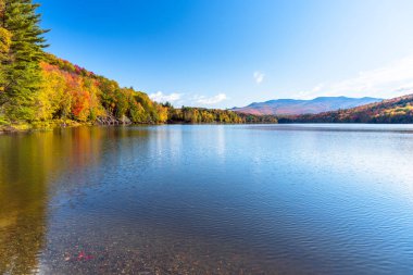 Beautiful mountain lake surrounded by deciduous woodland at the peak of fall foliage on a clear morning. Reflection in water. Waterbury, VT, USA. clipart