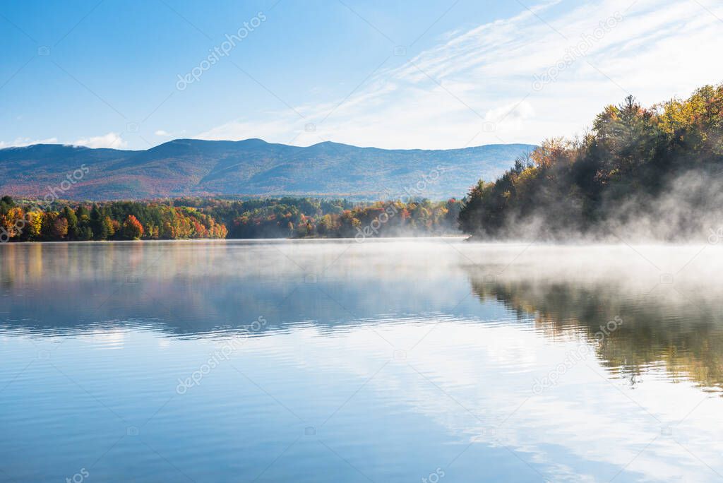 Majestic mountain lake with forested shores in autumn. The surface of the lake is covered in morning fog. Stunning autumn colours. Waterbury, VT, USA.