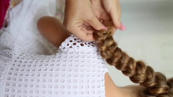 Вертикальное видео, где мать заплетает своим дочерям длинные светлые волосы. Кавказские женские руки. Маленькая девочка в белом платье. Вид сзади без лица — стоковое видео