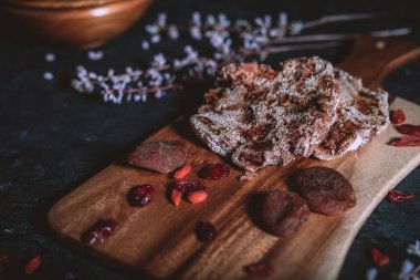 Ahşap kesim tahtasındaki ev yapımı meyve ekmeği dağınık kuru meyveler, çiçekler ve arka plandaki ahşap kâse siyah masanın üzerinde.