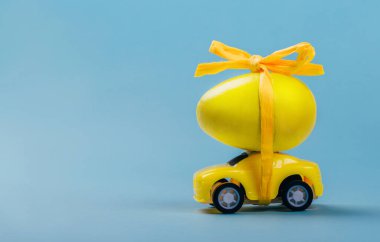 Yellow car toy with colored egg on top tied by string with bow on blue background.
