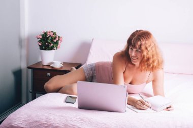 Evde dizüstü bilgisayarla çalışan orta yaşlı mutlu bir kadın evdeki yatakta oturuyor..