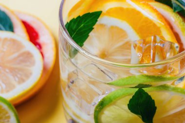 Limonlu, limonlu, portakallı ve greyfurtlu dilimler, nane yapraklı ve buz küplü bir bardak suda ve sarı arka planda..