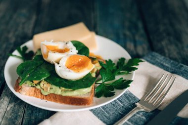 Avokadolu tost, haşlanmış yumurta ıspanak ve maydanozlu beyaz tabak üzerinde bıçak ve çatallı peçete ve kırsal mavi ahşap arka plan.