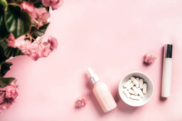 Botol kosmetik dan kapsul putih dengan bunga begonia di latar belakang kertas merah muda. Konsep kesehatan dan kecantikan — Stok Foto