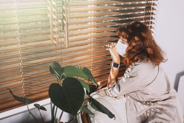 Orta yaşlı kadın maske takıyor ve perdelerden dışarı bakıyor. Karantinadaki kadın pencereden bakıyor..