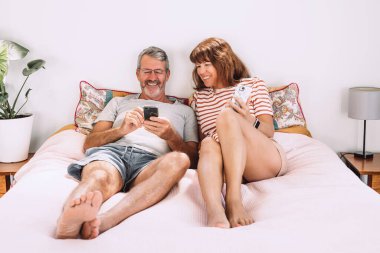 Orta yaşlı çift cep telefonlarını kullanıyor. Yatakta uzanıyor ve gülüyorlar..