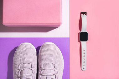 Yeni mor spor ayakkabılar, pembe yoga bloğu ve renkli arka planda akıllı saat. Koşu ayakkabılarının yukarıdan çekimi.