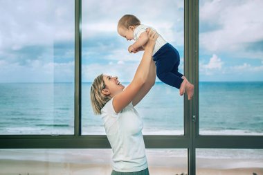 Bir anne küçük bebeğini kucağına alır ve güzel bir okyanus manzarası olan büyük pencerenin yanında kahkaha atar.