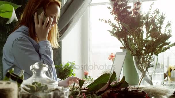 Blomsterhandler som arbeider med bærbar datamaskin og snakker per telefon – stockvideo