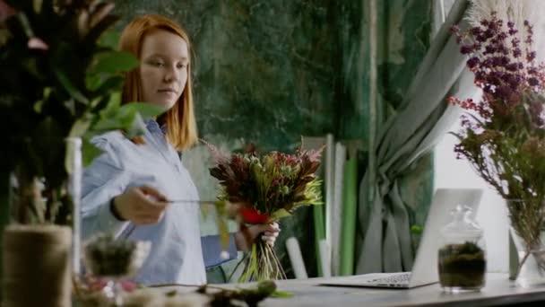 Fleuriste gingembre créant un bouquet de fleurs près d'un ordinateur portable sur la table — Video