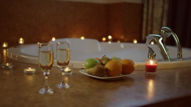 Assiette avec des fruits et des verres de champagne pour deux debout près de l'eau coulant dans le bain — Video