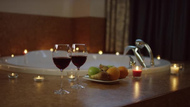 Assiette avec des fruits et des verres de vin rouge pour deux debout près de l'eau coulant dans le bain — Video