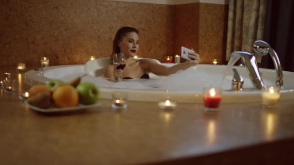 Жінка з червоним вином у ванні з піною посміхається на селфі — стокове відео