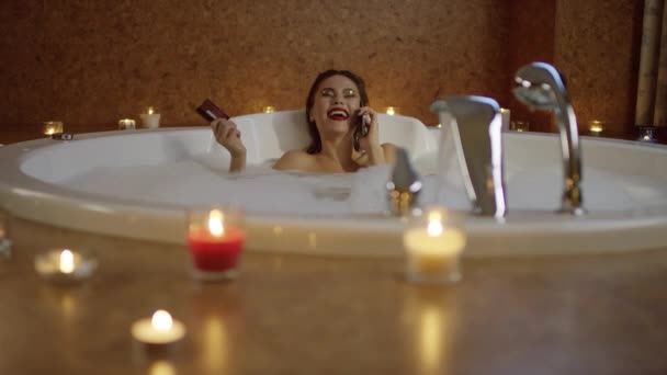 Mujer riendo en el baño con espuma hablando por teléfono y la celebración de la tarjeta bancaria . — Vídeos de Stock