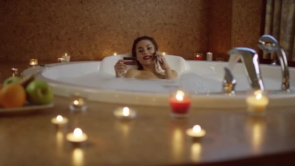 Femme dans le bain avec de la mousse parlant au téléphone et tenant la carte bancaire et rire — Video