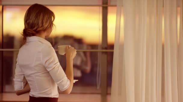 Genç kadın pencerenin önünde duruyor ve bir fincan çay ya da kahve içiyor — Stok video