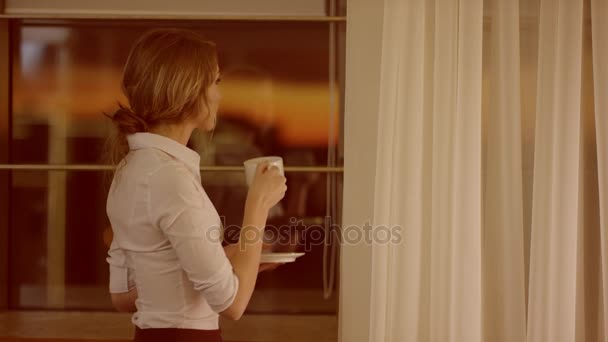 Jovem fica na frente da janela e bebe uma xícara de chá ou café — Vídeo de Stock