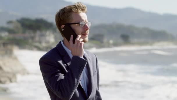 Young businessman stands by the Pacific ocean coast and talks on phone — Stock Video